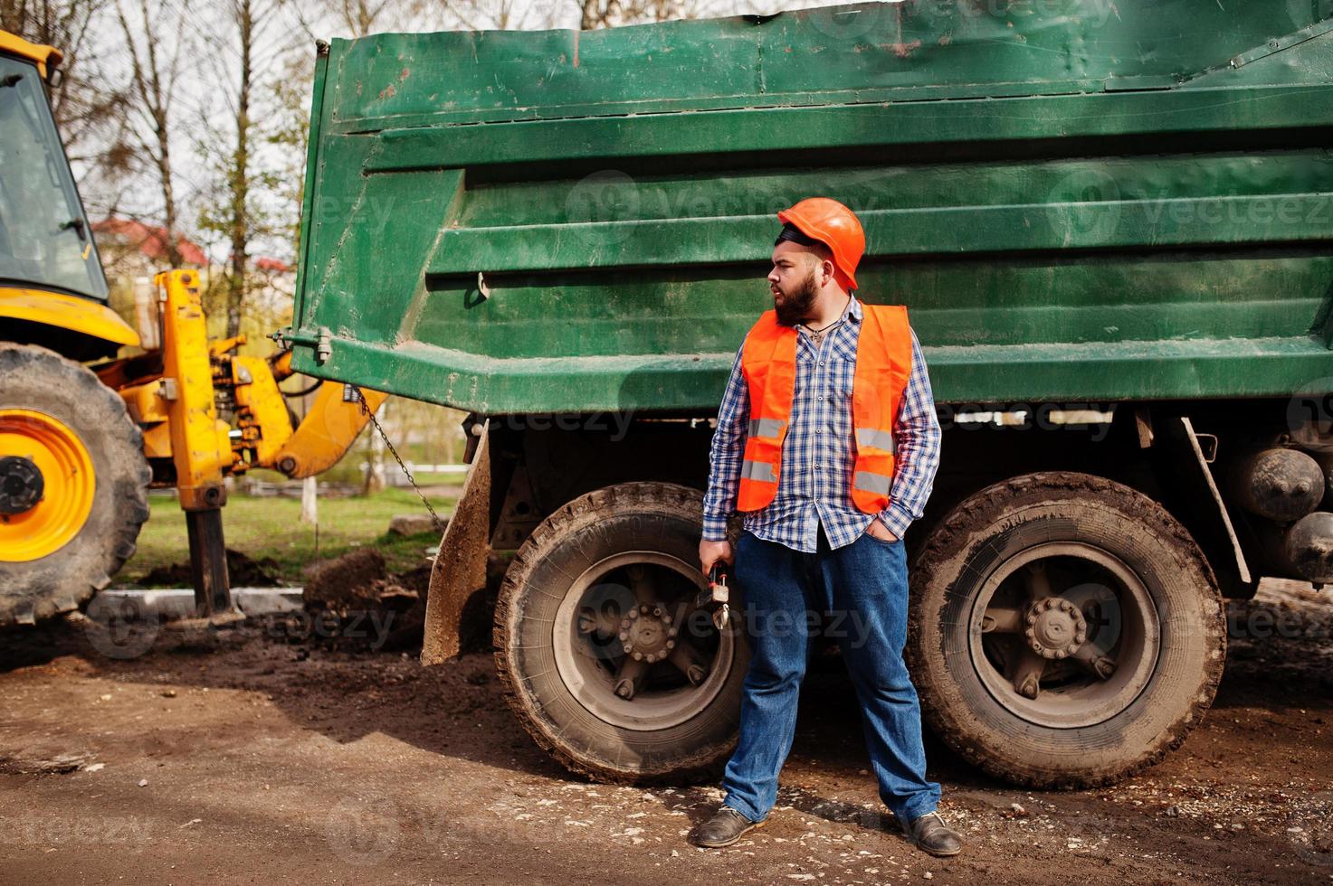 Brutal beard worker man suit construction worker in safety orange helmet, against dump truck with hammer and adjustable wrench at hand. photo