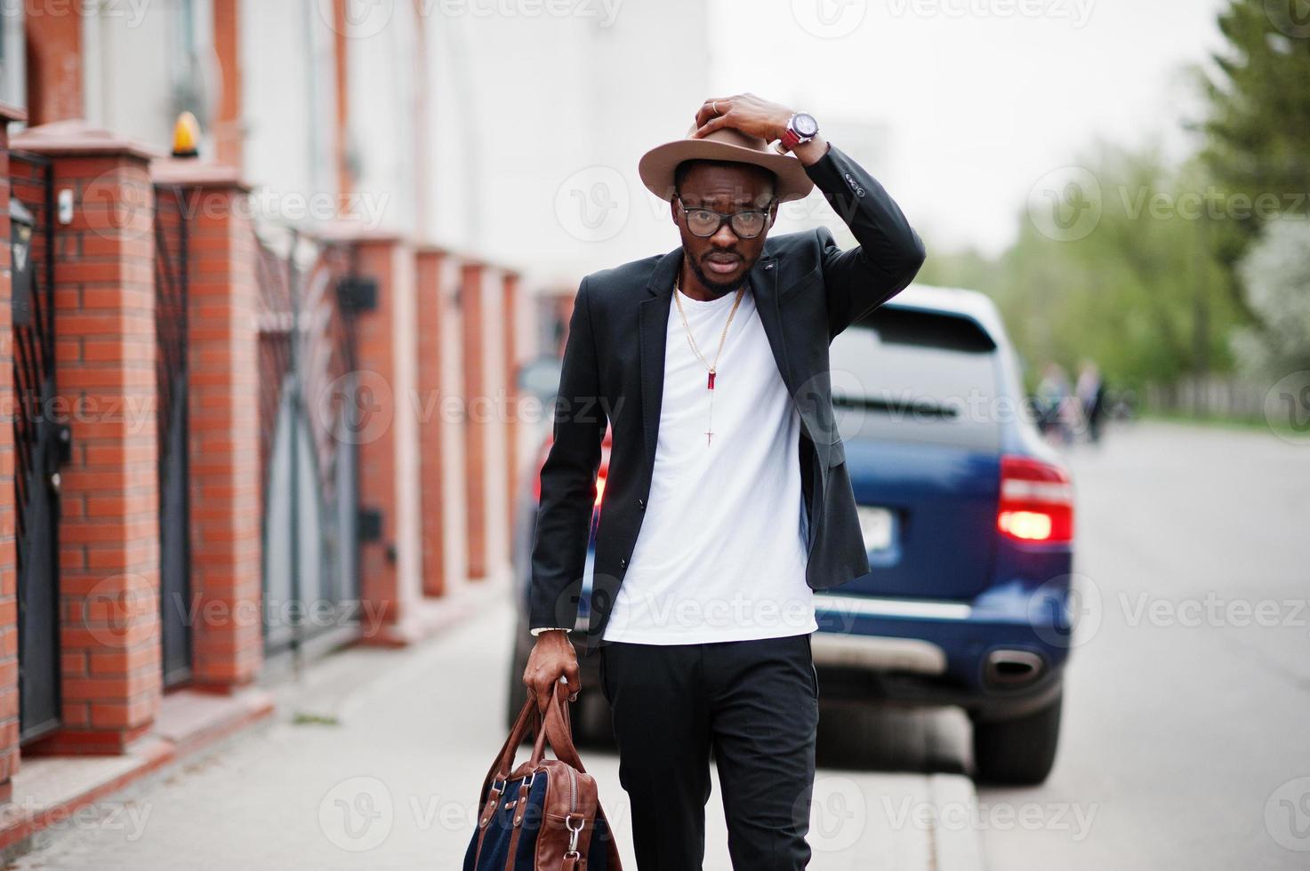 hombre negro con estilo en gafas con sombrero, vestido con traje con bolso contra coche de lujo. rico hombre de negocios afroamericano. foto