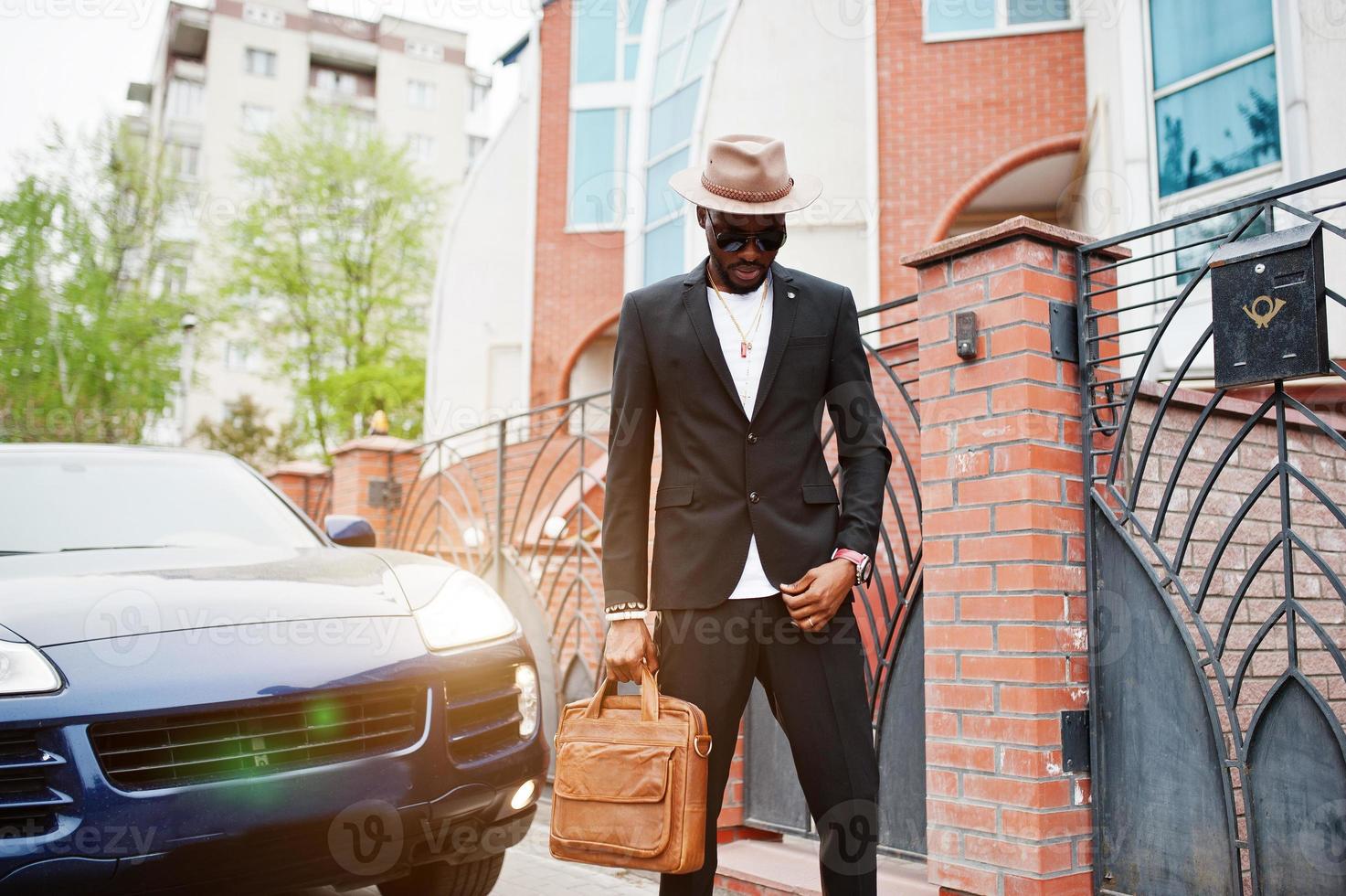 hombre negro con estilo en gafas con sombrero, vestido con traje con bolso contra coche de lujo. rico hombre de negocios afroamericano. foto