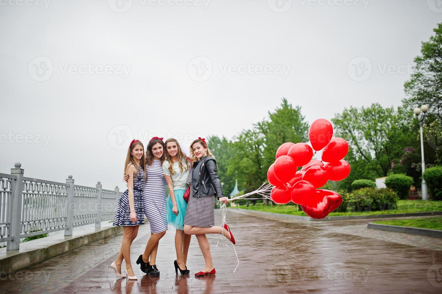 Attractive bride posing with her three lovely bridesmaids with red heart-shaped balloons on the pavement with lake on the background. Bachelorette party. photo