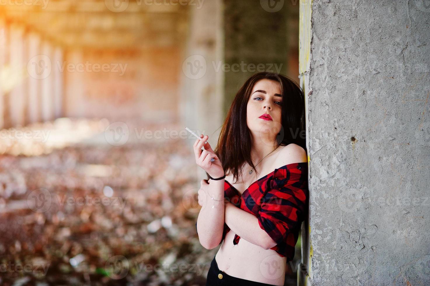 Retrato de una chica fumadora con labios rojos que llevaba una camisa a cuadros roja con los hombros desnudos posó un lugar sexy de fondo abandonado. foto