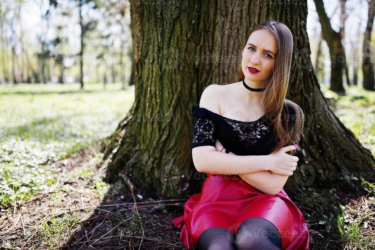 retrato de una chica con maquillaje brillante con labios rojos, collar de gargantilla negra en el cuello y falda de cuero rojo sentada cerca de un árbol en el parque. foto