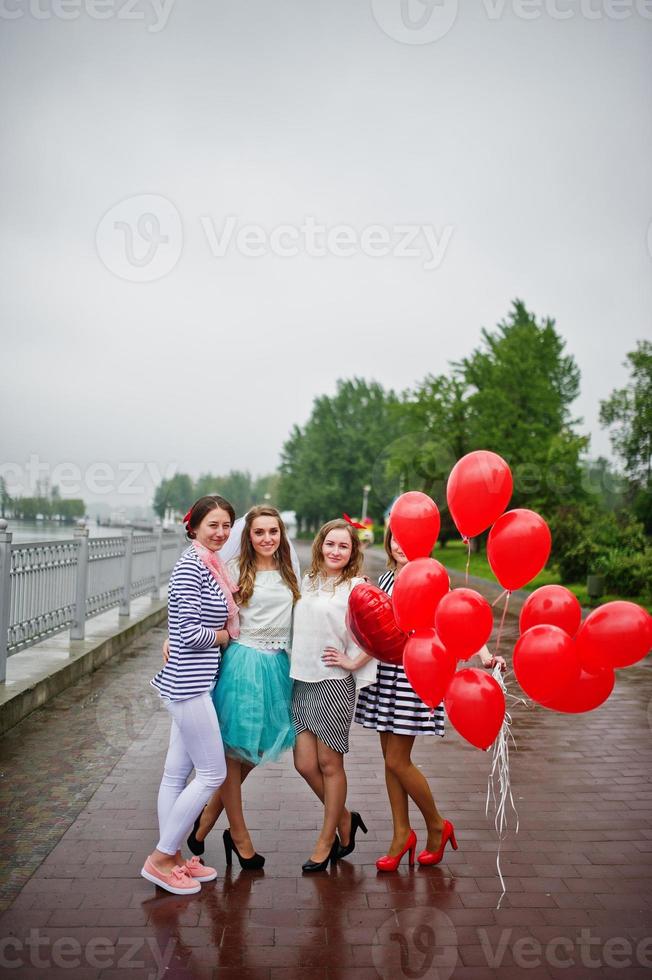 atractiva novia posando con sus tres encantadoras damas de honor con globos rojos en forma de corazón en el pavimento con un lago al fondo. despedida de soltera. foto