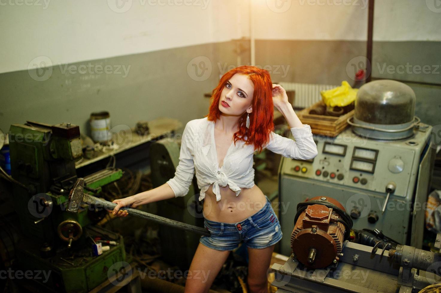 la chica de cabello rojo usa pantalones cortos de mezclilla y blusa blanca con un martillo en las manos posado en una máquina industrial en la fábrica. foto