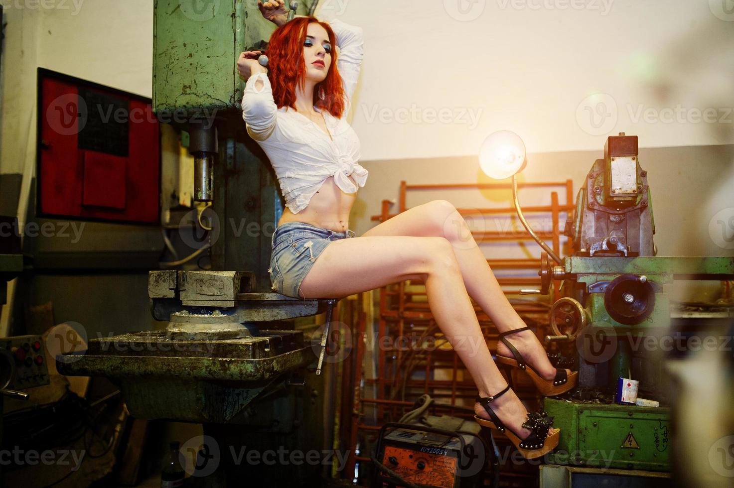 Red haired girl wear on short denim shorts and white blouse posed at industrial machine at the factory. photo