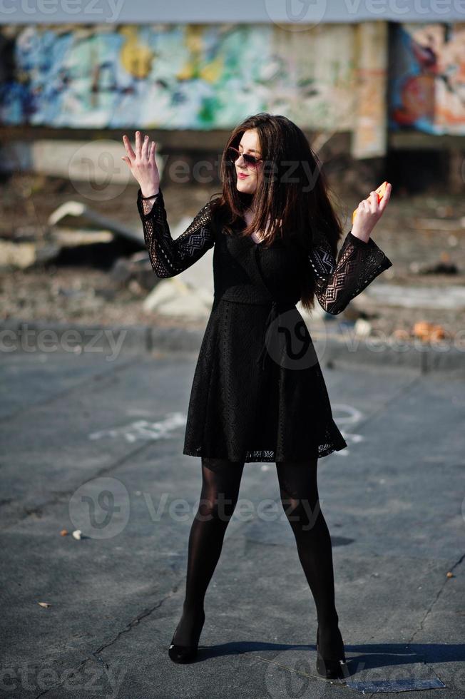 Portrait brunette girl with red lips and orange mobile phone at hands, wearing a black dress, sunglasses posed on the roof. Street fashion model. photo