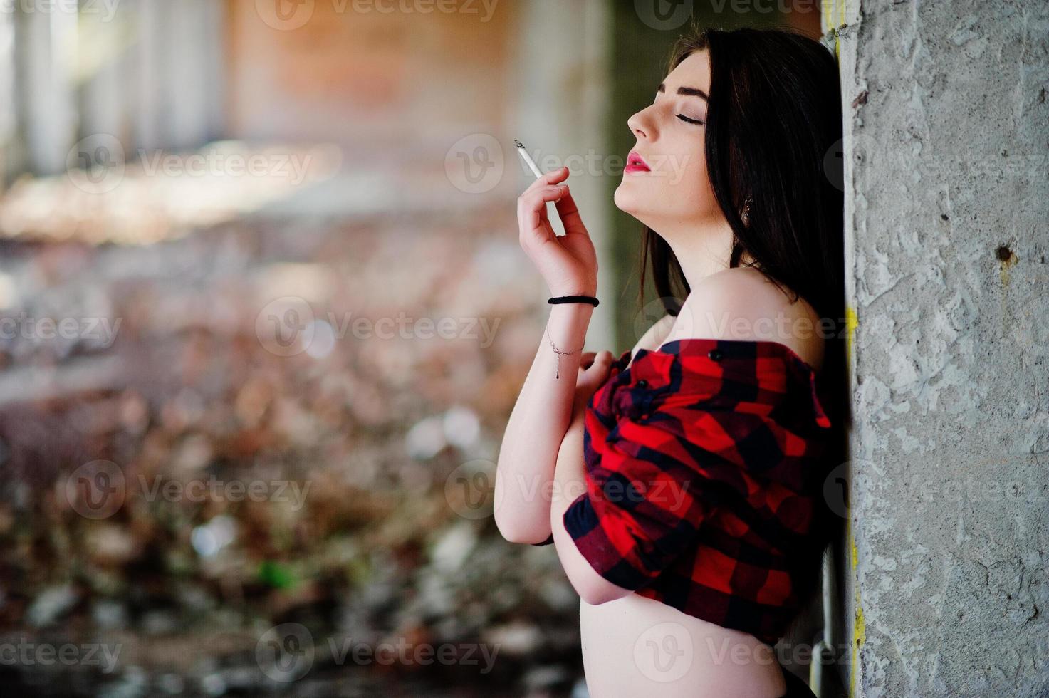 Retrato de una chica fumadora con labios rojos que llevaba una camisa a cuadros roja con los hombros desnudos posó un lugar sexy de fondo abandonado. foto