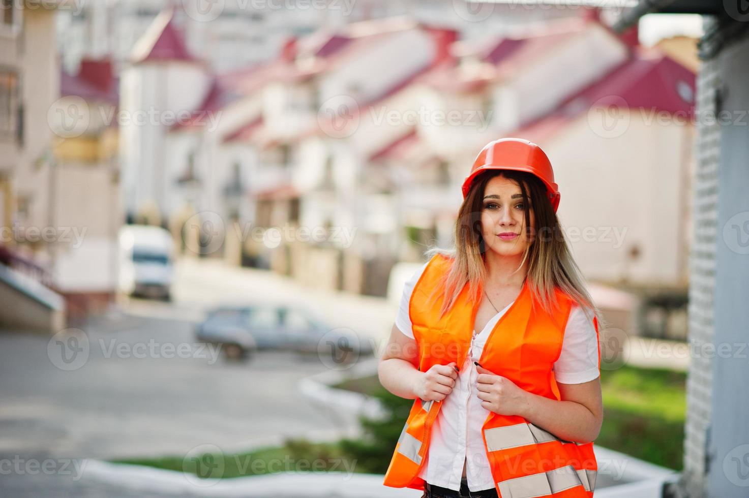 Engineer builder woman in uniform waistcoat and orange protective helmet against new building. Property living block theme. photo