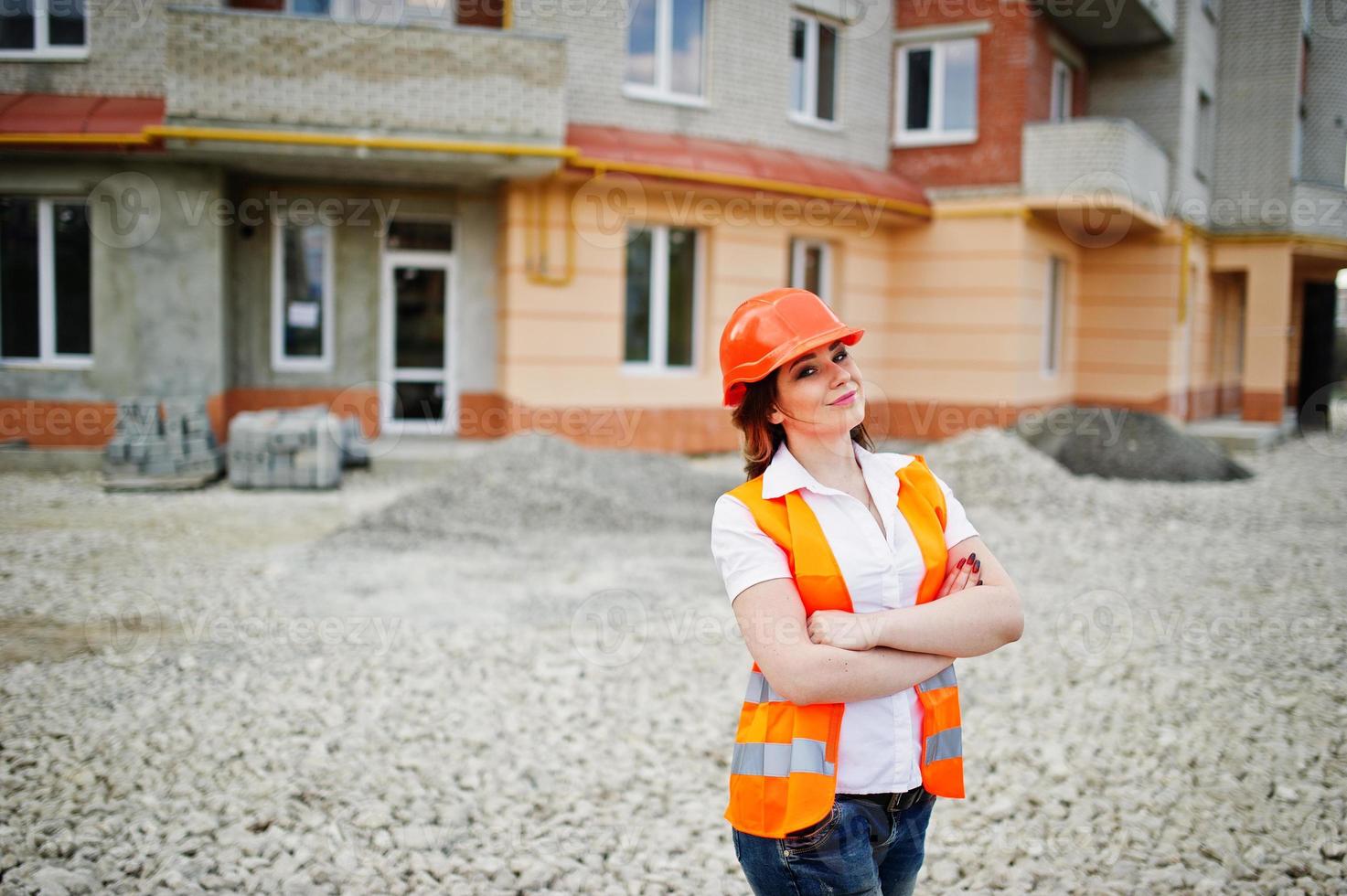 Engineer builder woman in uniform waistcoat and orange protective helmet against new building. Property living block theme. photo