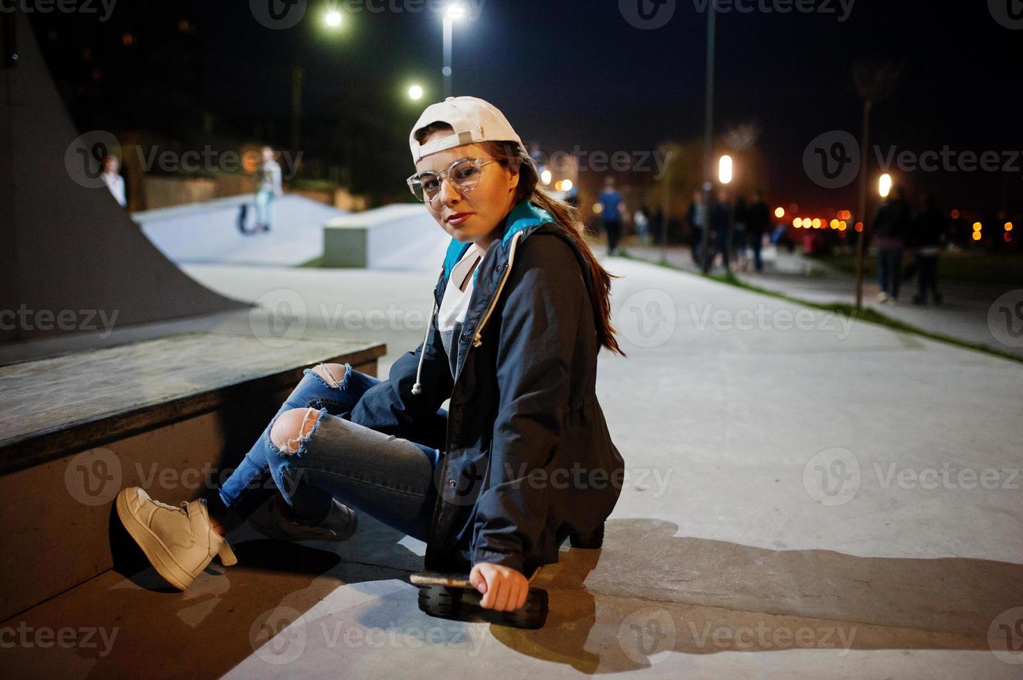 Young teenage urban girl with skateboard, wear on glasses, cap and ripped jeans at skate park on the evening. photo