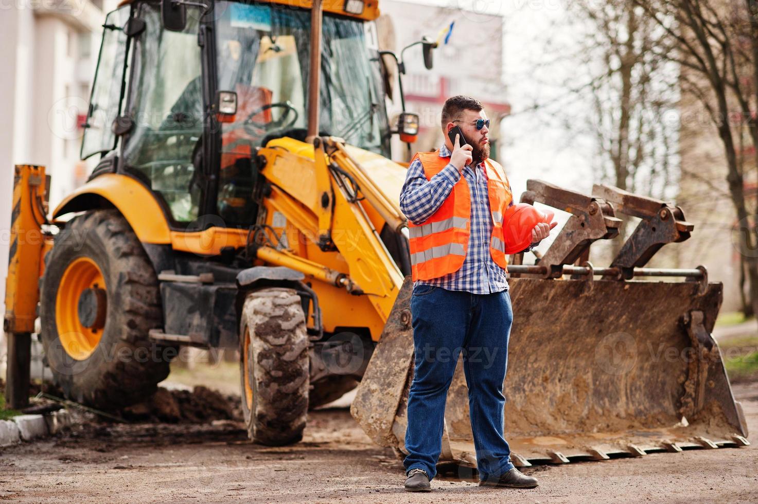 trabajador de barba traje de hombre trabajador de la construcción en casco naranja de seguridad, gafas de sol contra tractor con teléfono móvil a mano. foto