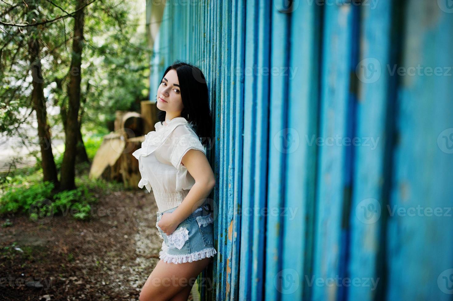 retrato de chica morena sexy en shorts de jeans de mujer y blusa blanca contra casa de madera azul con tocones. foto