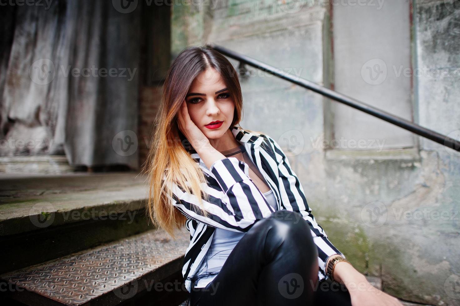 mirada de mujer de moda con chaqueta de traje a rayas blancas y negras, pantalones de cuero, posando en la calle vieja en escaleras de hierro. concepto de chica de moda. foto