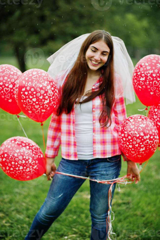 Portrait of brunette girl on checkered shirt, jeans and veil with many red balloons at hen party. photo