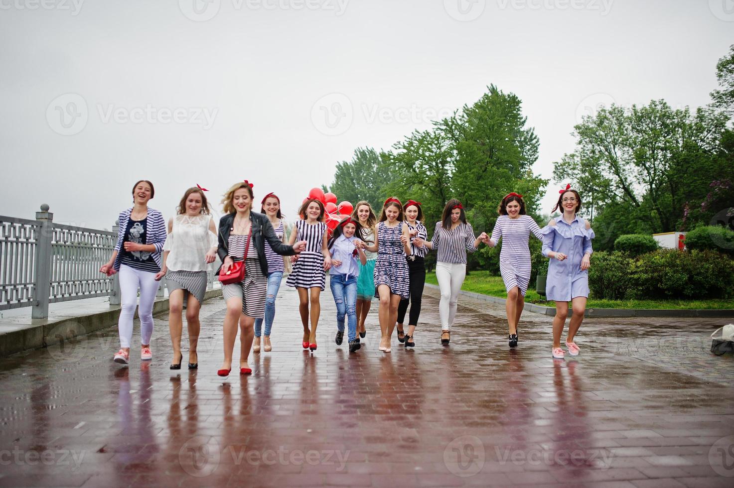 Eleven amazingly-looking braidsmaids with stunning bride posing with red heart-shaped balloons on the pavement against the lake in the background. photo