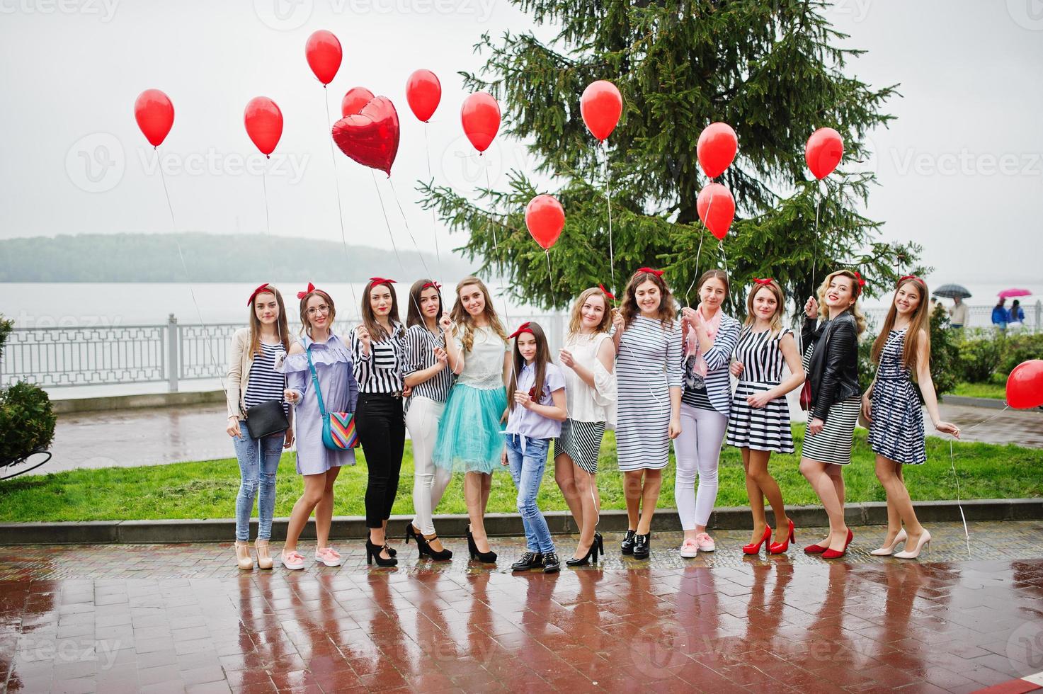 Eleven amazingly-looking braidsmaids with stunning bride posing with red heart-shaped balloons on the pavement against the lake in the background. photo