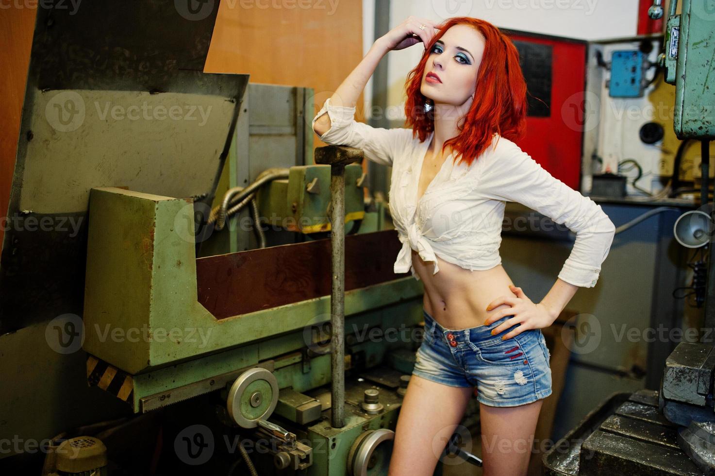 Red haired girl wear on short denim shorts and white blouse with hammer at hands posed at industrial machine at the factory. photo