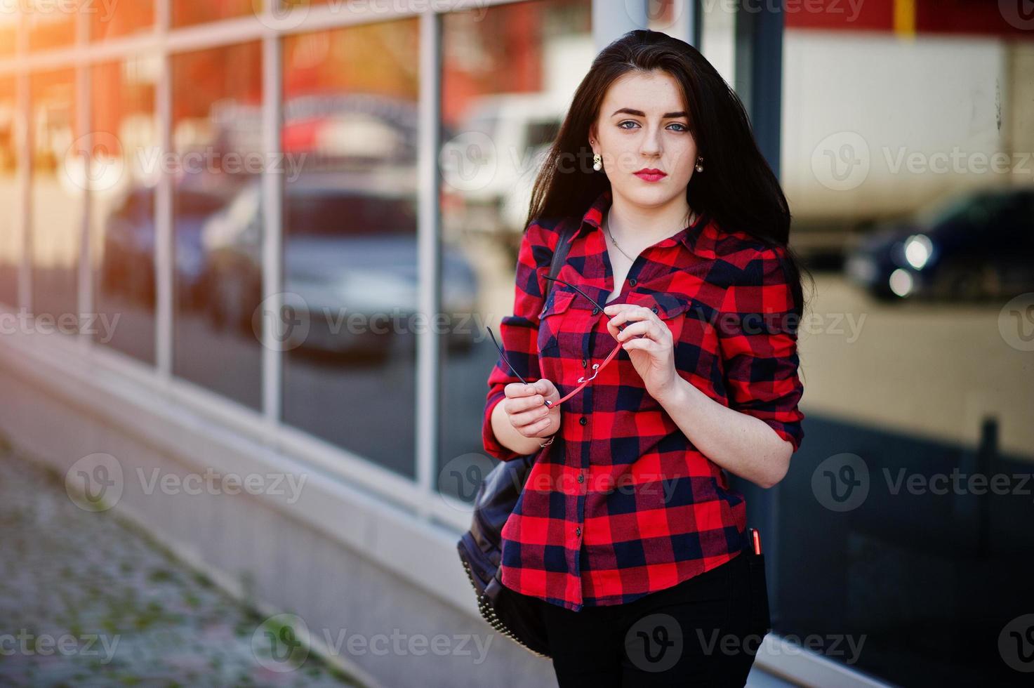 Fashion portrait girl with red lips wearing a red checkered shirt and backpack holding sunglasses at hands background large mirror windows. photo