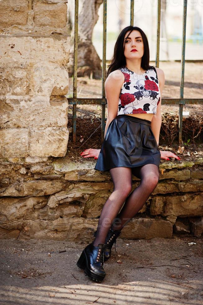 Young goth girl on black leather skirt and high heels punk shoes posed against iron fence. photo