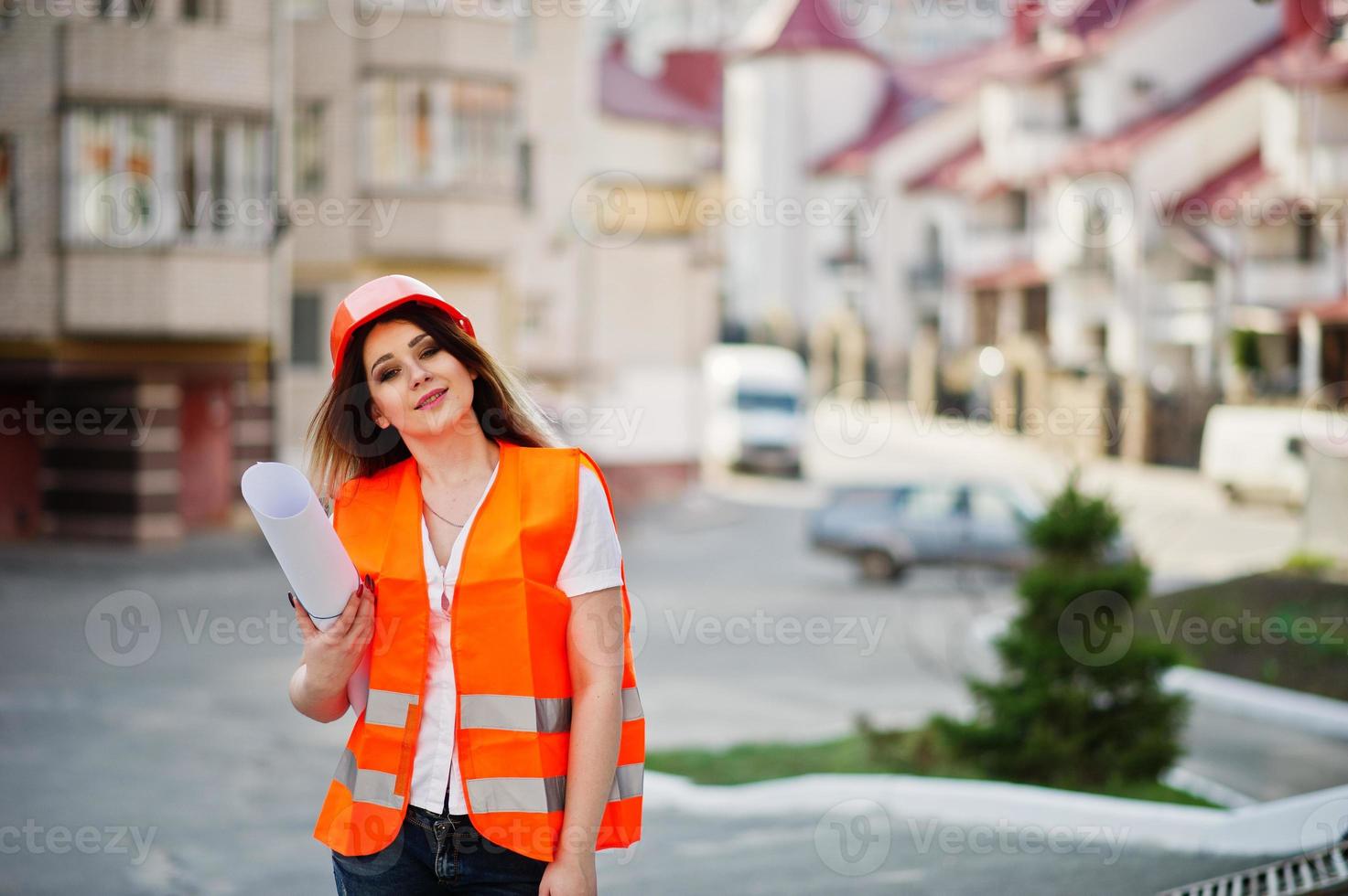 Engineer builder woman in uniform waistcoat and orange protective helmet hold business drawing paper roll against new building. Property living block theme. photo