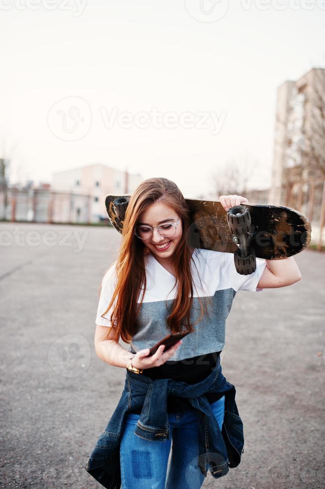 Young teenage urban girl with skateboard, wear on glasses, cap and ripped jeans at the yard sports ground on sunset making selfie on phone. photo