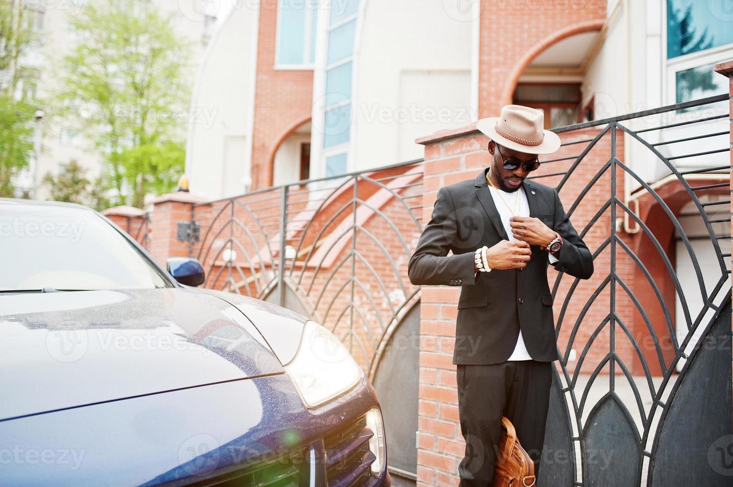 hombre negro con estilo en gafas con sombrero, vestido con traje con bolso contra coche de lujo. rico hombre de negocios afroamericano. foto