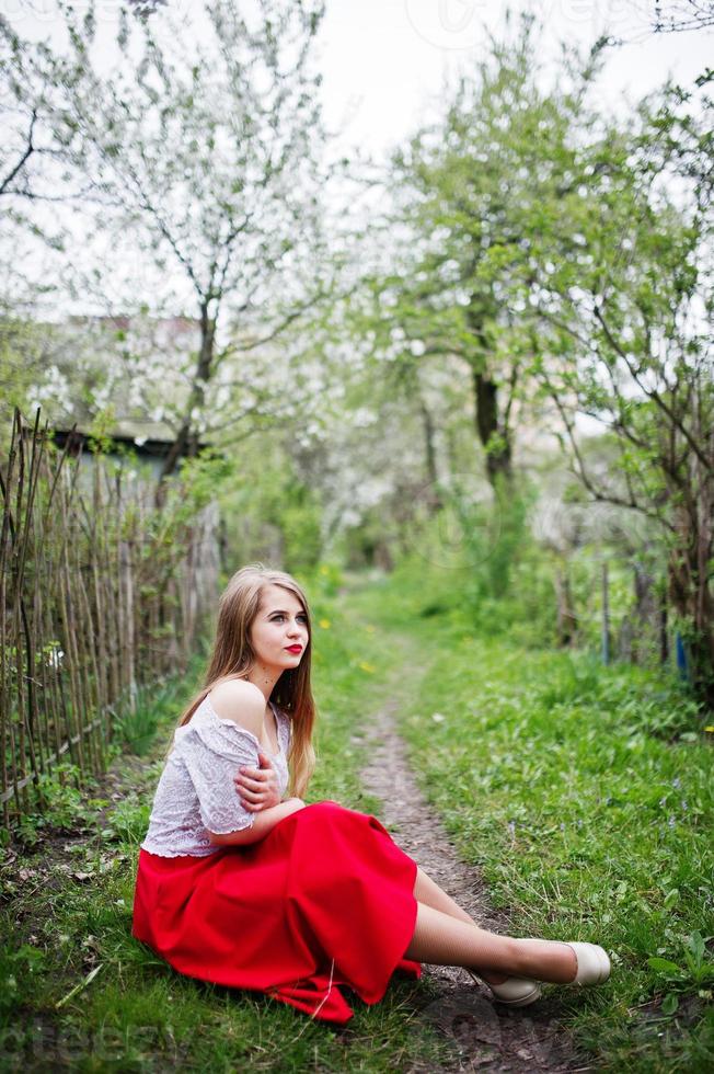 Portrait of sitting beautiful girl with red lips at spring blossom garden on green grass, wear on red dress and white blouse. photo