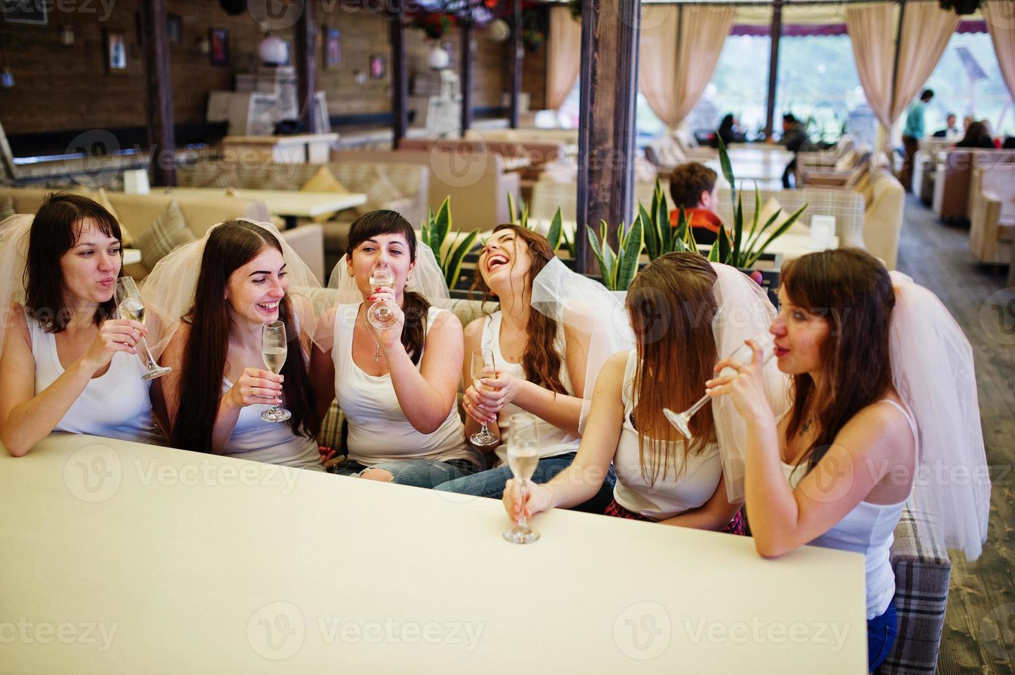 Group of cheerful girls at white shirts sitting at table and drink champagne on hen party. photo