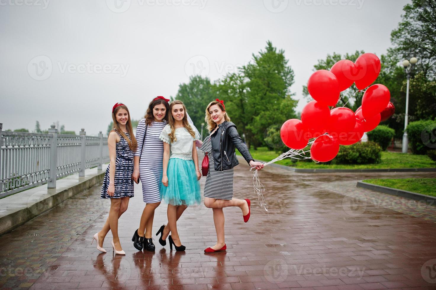 Attractive bride posing with her three lovely bridesmaids with red heart-shaped balloons on the pavement with lake on the background. Bachelorette party. photo