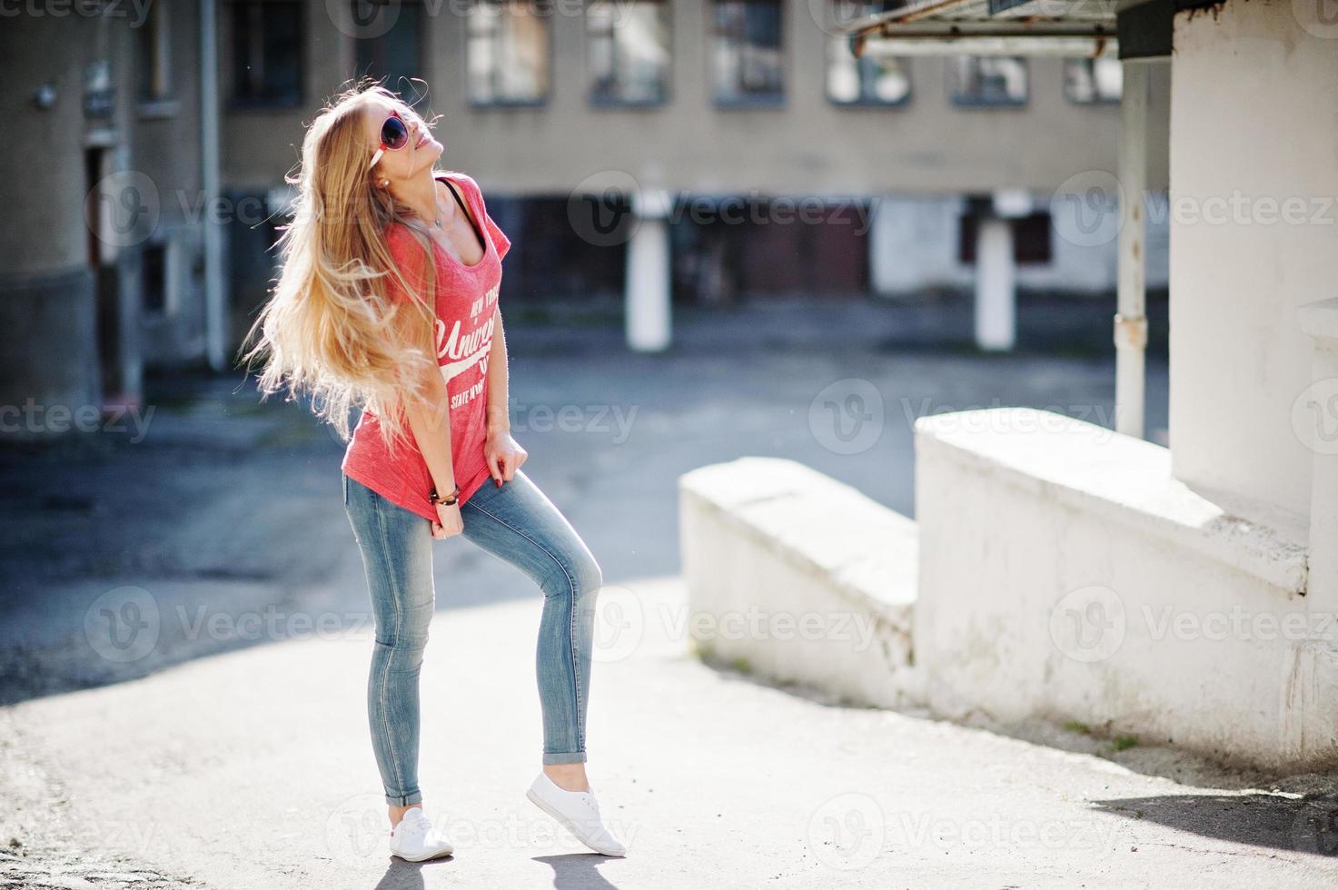 Stylish happy blonde woman wear at jeans, sunglasses and t-shirt posed at street on sunny weather. Fashion urban model portrait. photo