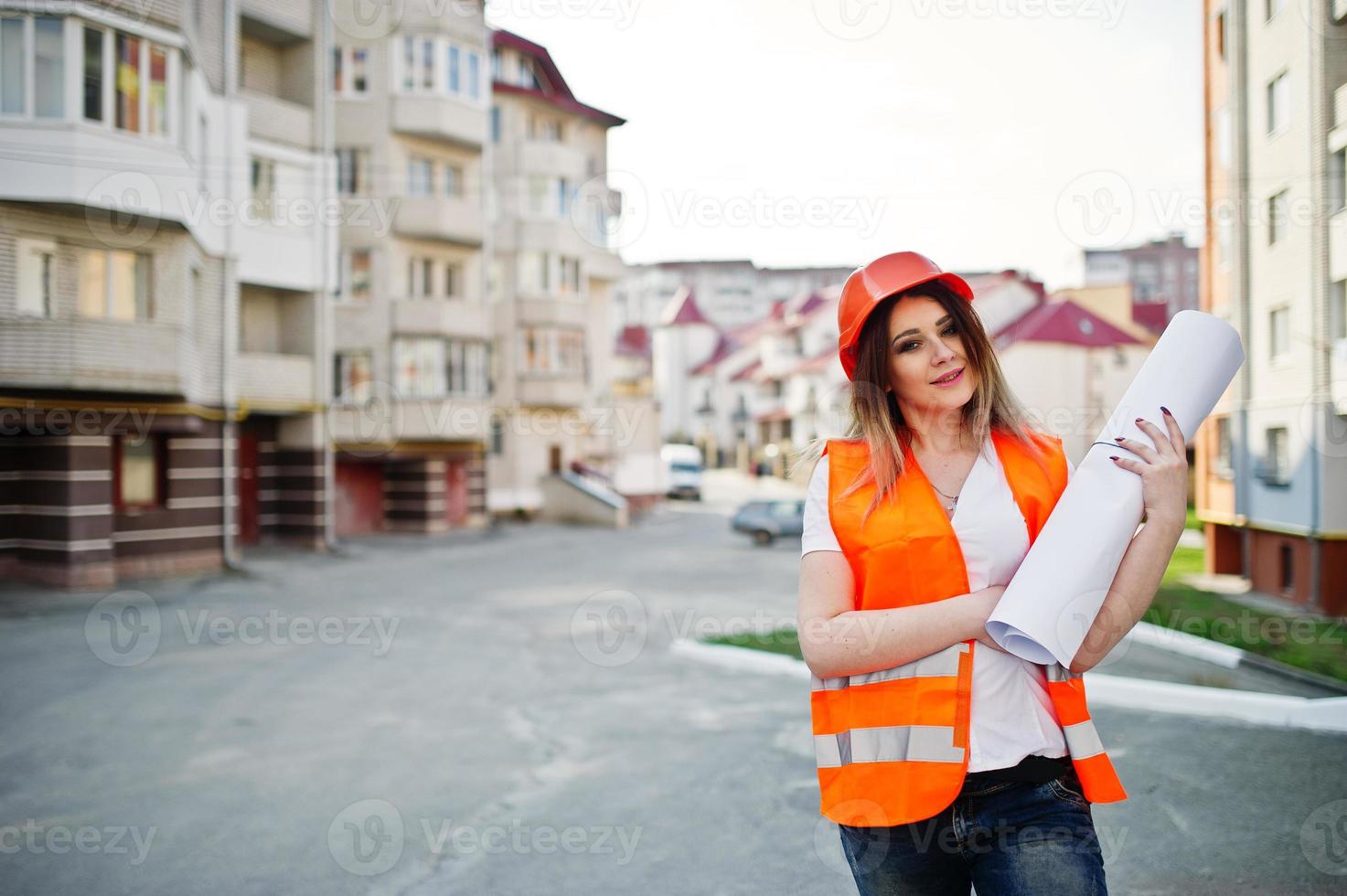 mujer ingeniera constructora con chaleco uniforme y casco protector naranja sostiene un rollo de papel de dibujo comercial contra un nuevo edificio. tema de bloque de vivienda de propiedad. foto