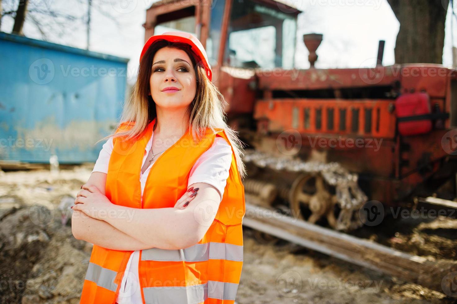 Engineer builder woman in uniform waistcoat and orange protective helmet against bulldozer. photo