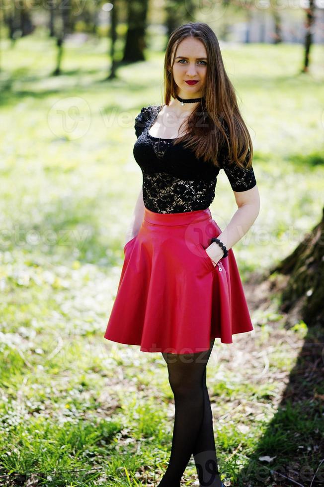 retrato de niña con maquillaje brillante con labios rojos, collar de gargantilla negra en el cuello y falda de cuero rojo en el parque de primavera. foto