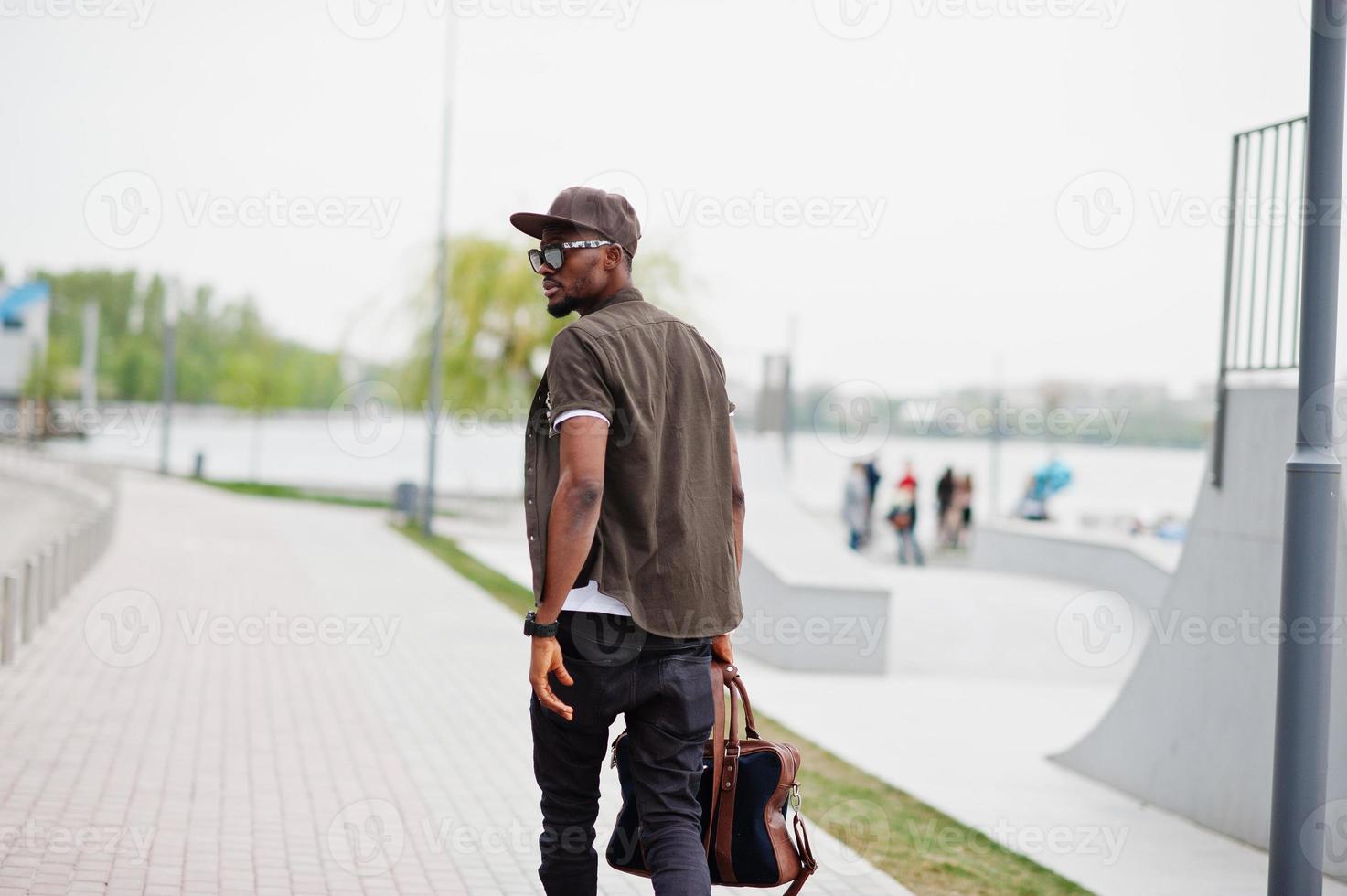 retrato de vista trasera de un hombre afroamericano elegante que camina con gafas de sol y gorra con bolso al aire libre. hombre negro de moda callejera. foto