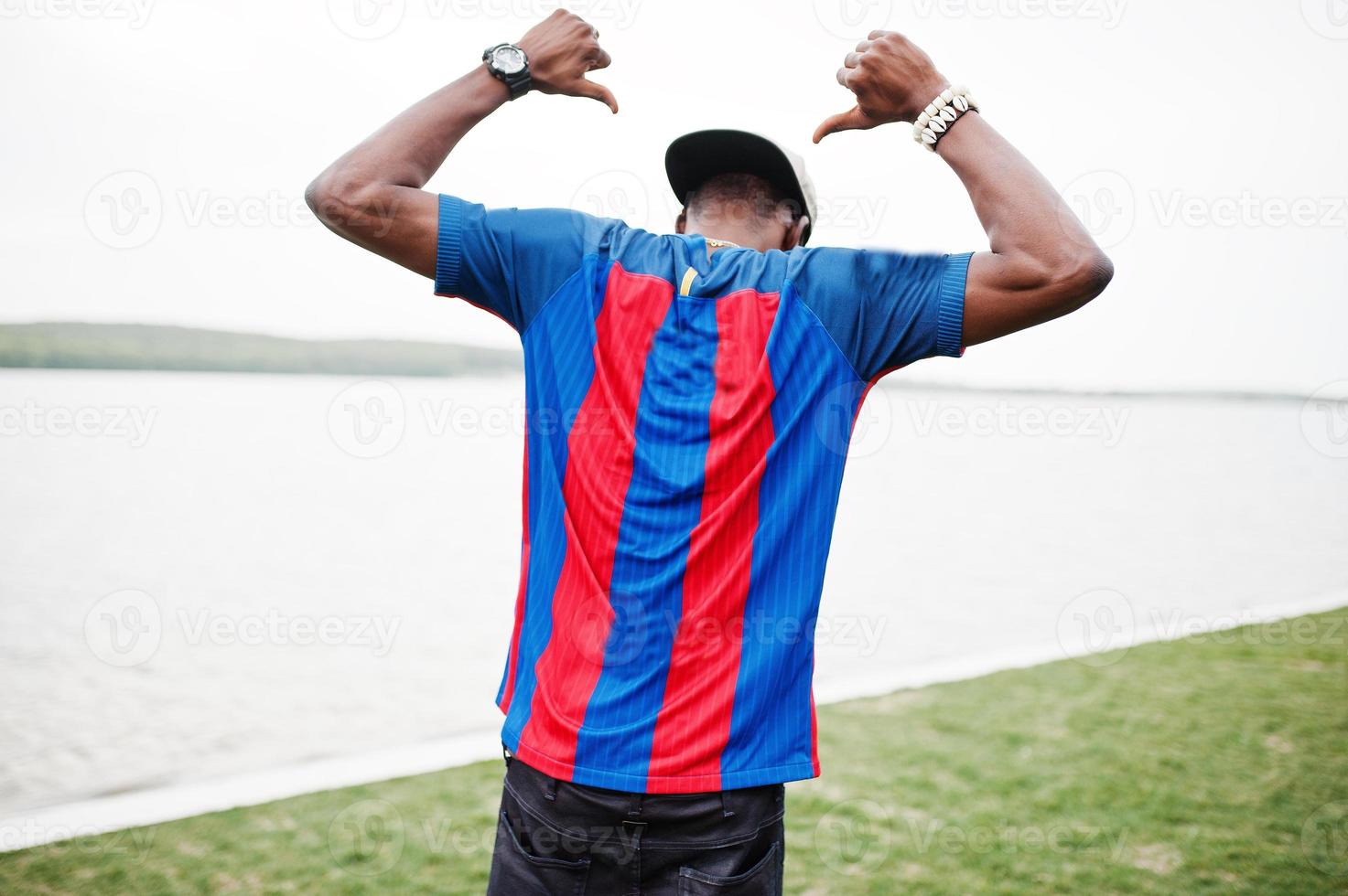 Stylish african american boy wear at cap, football t-shirt showing his back against lake. Black sports man portrait. photo