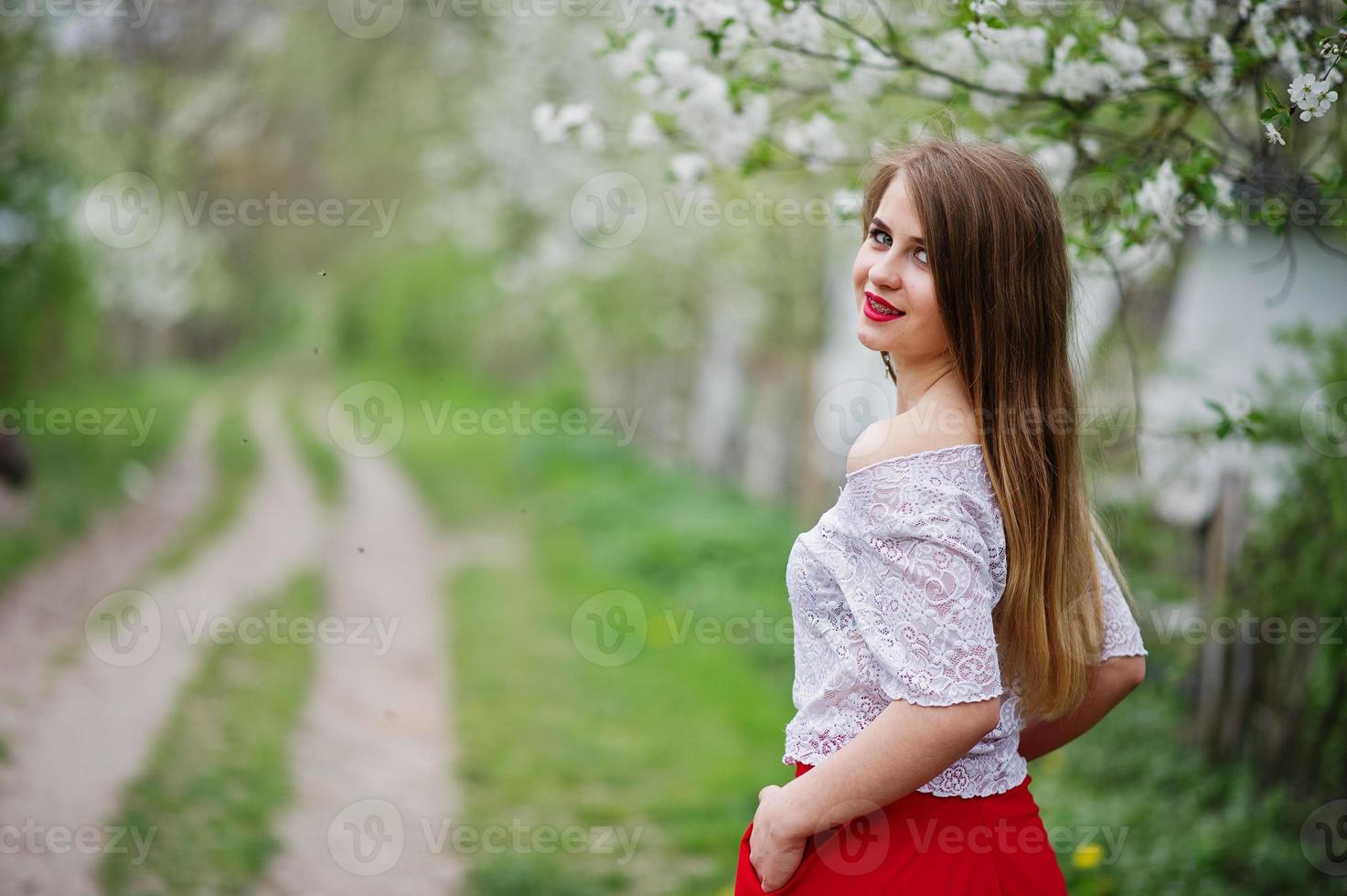 retrato de una hermosa chica con labios rojos en el jardín de flores de primavera, vestido rojo y blusa blanca. foto