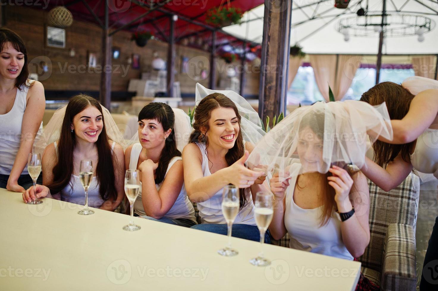 Group of cheerful girls at white shirts sitting at table and drink champagne on hen party. photo