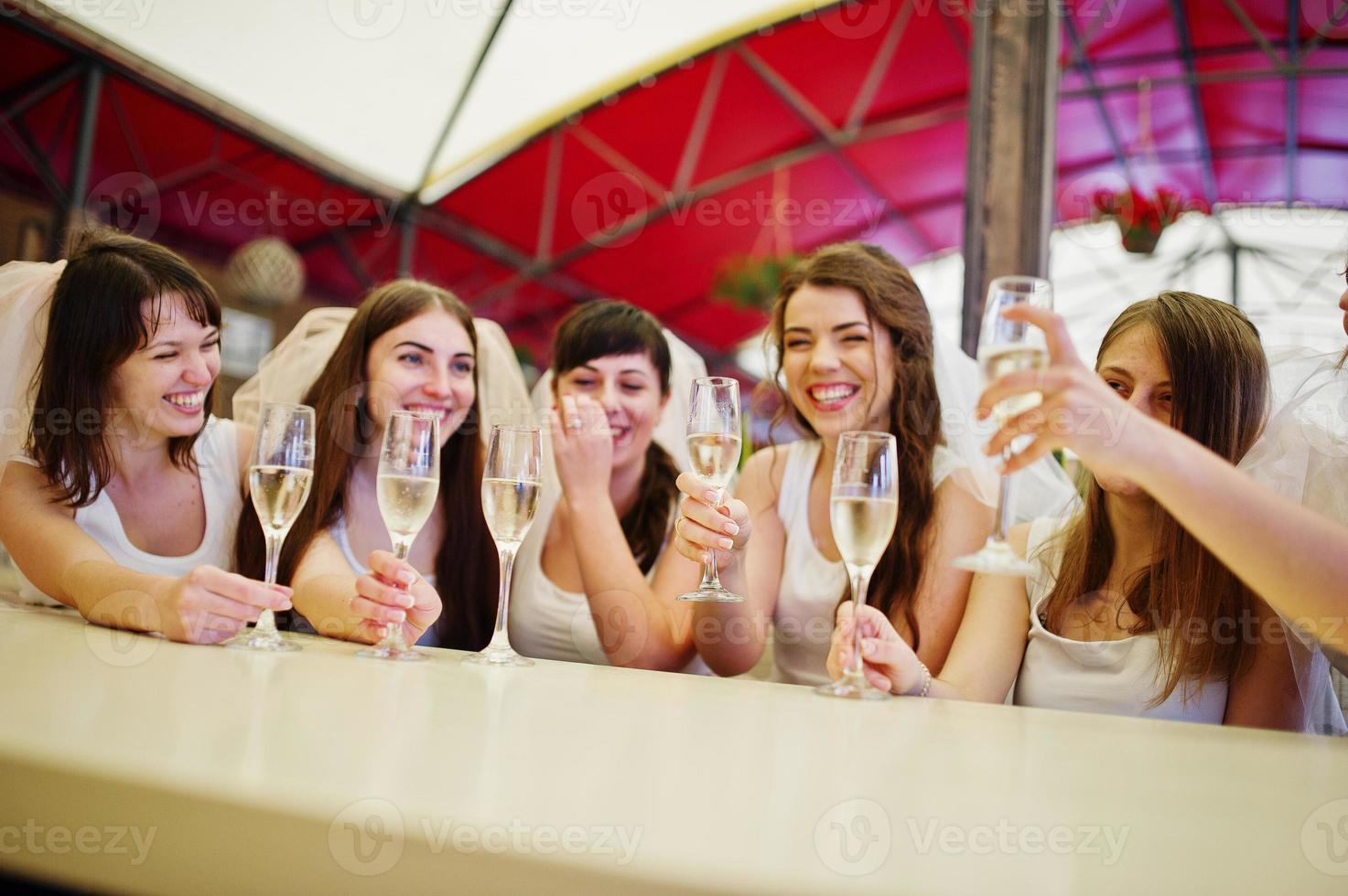 grupo de chicas alegres con camisas blancas sentadas en la mesa y bebiendo champán en la despedida de soltera. foto