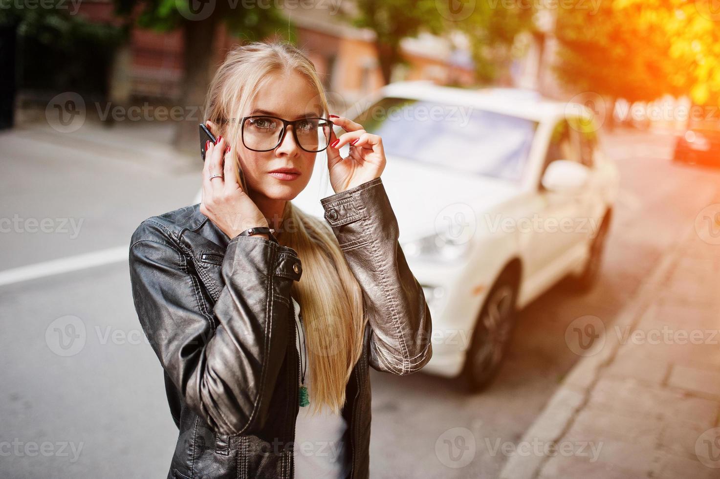 Elegante mujer rubia vestida con jeans, gafas y chaqueta de cuero con teléfono móvil, contra un auto de lujo. retrato de modelo urbano de moda. foto