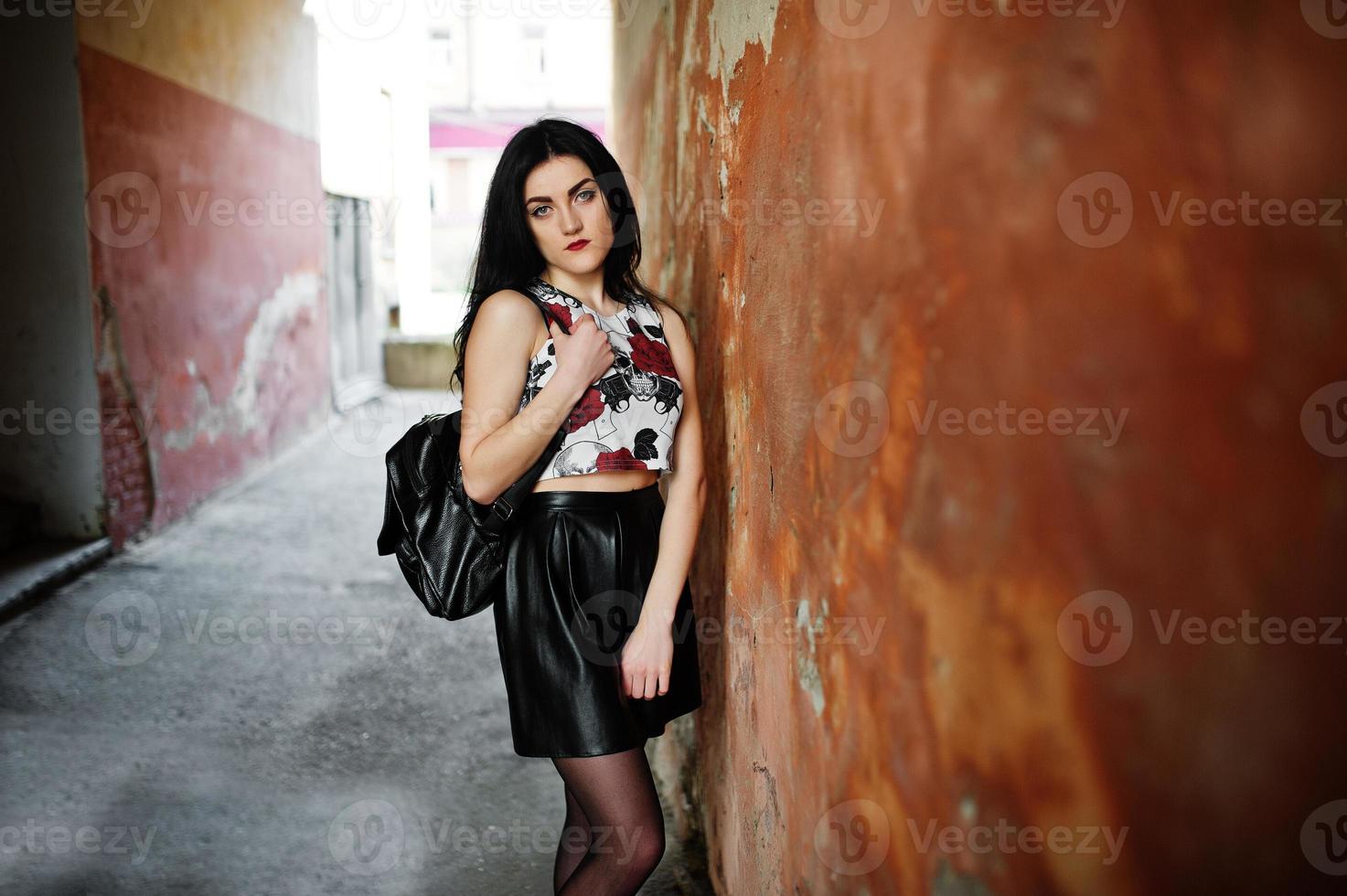 joven gótica en falda de cuero negro con mochila posada contra la pared del grunge. foto