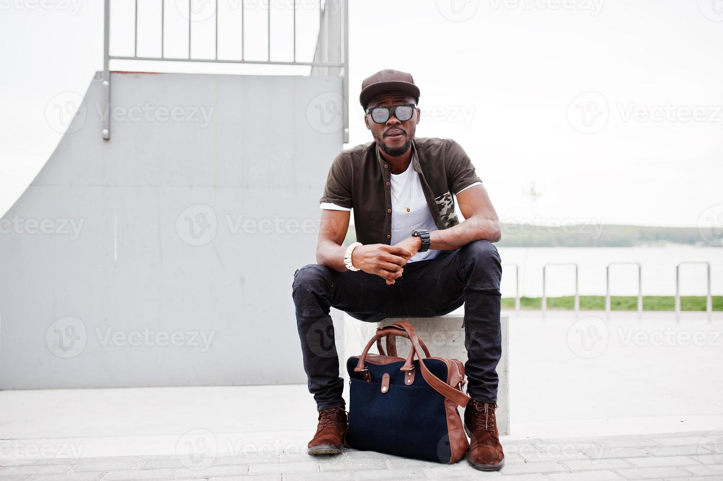 Portrait of sitting stylish african american man wear on sunglasses and cap, looking on his watches outdoor. Street fashion black man. photo