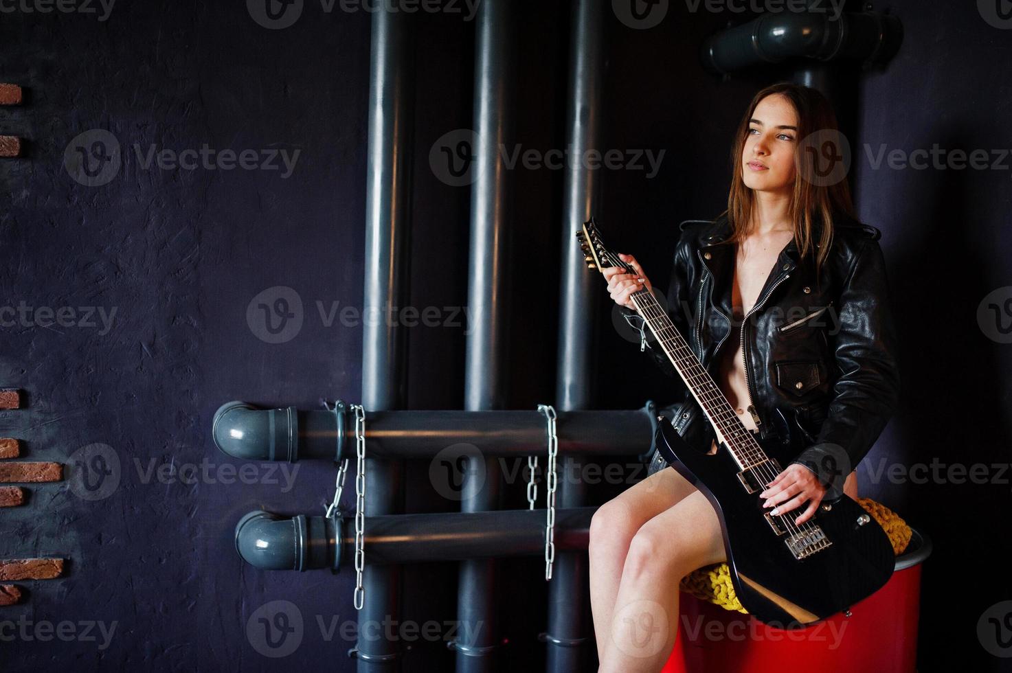 Portrait of sexy nude brunette girl at black leather jacket with guitar against industrial background with red barrel. Fashion model studio shoot. photo