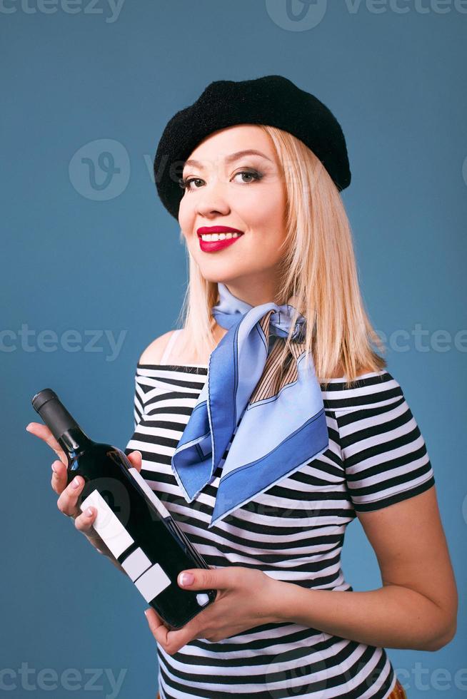 Retrato de una hermosa mujer francesa rubia en boina, bufanda, espalda y camisa blanca, con una botella de vino y pan baguette en sus brazos con la bandera francesa en el fondo foto