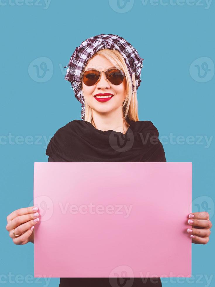 cute adorable cheerful smiling blonde woman in sunglasses and arafatka with pink empty table in her hands on blue background. Travel photo