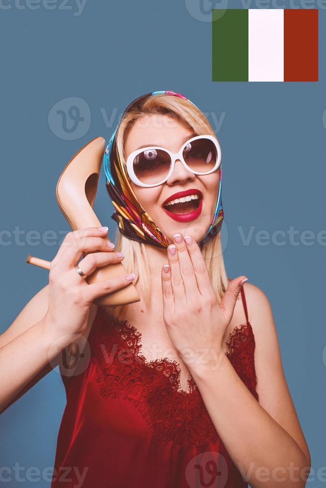 funny stylish smiling cheerful fancy blonde woman with make up and with high heel shoe in her arms and italian flag on background photo