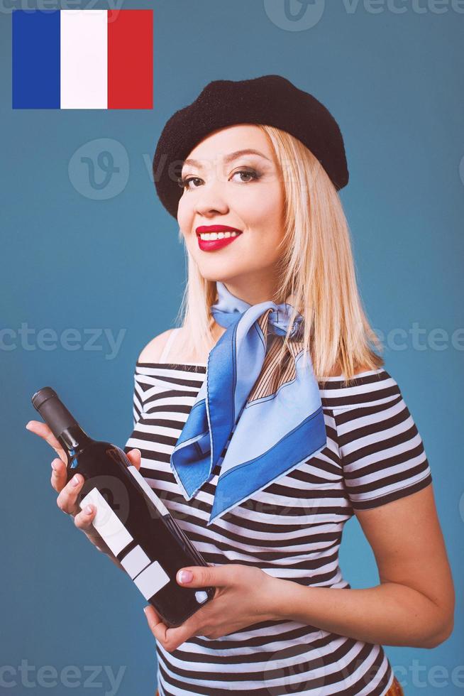 Retrato de una hermosa mujer francesa rubia en boina, bufanda, espalda y camisa blanca, con una botella de vino y pan baguette en sus brazos con la bandera francesa en el fondo foto