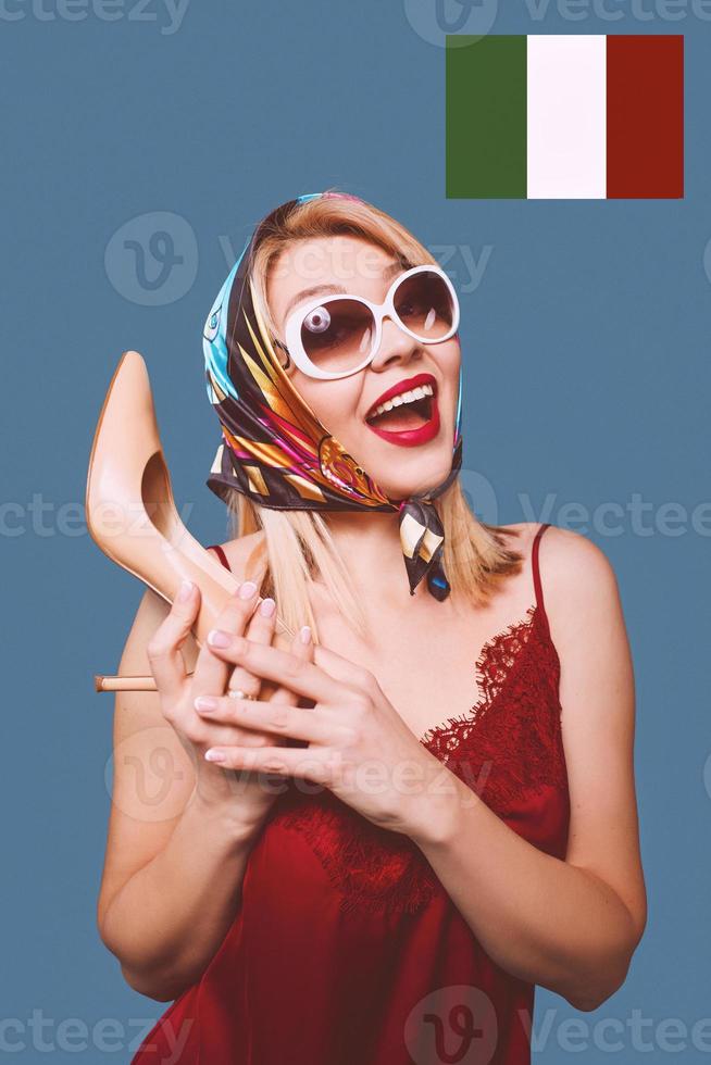 funny stylish smiling cheerful fancy blonde woman with make up and with high heel shoe in her arms and italian flag on background photo