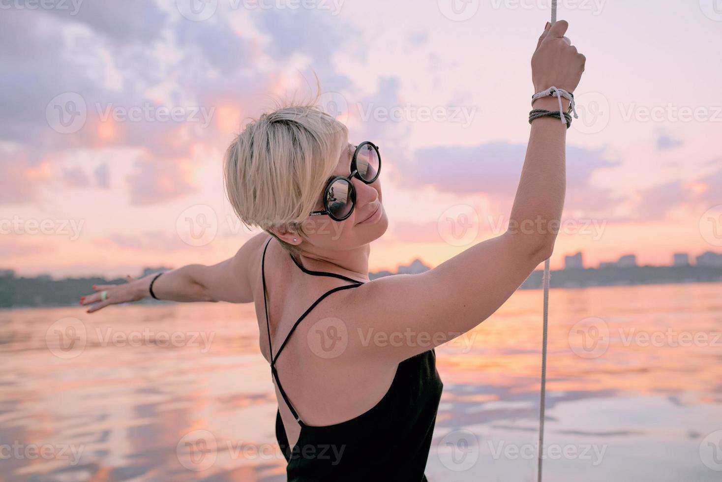 hermosa mujer rubia feliz con gafas de sol y vestido largo negro disfrutando de la puesta de sol en el yate en el mar. concepto de viaje, aventura, relajación, relajación foto