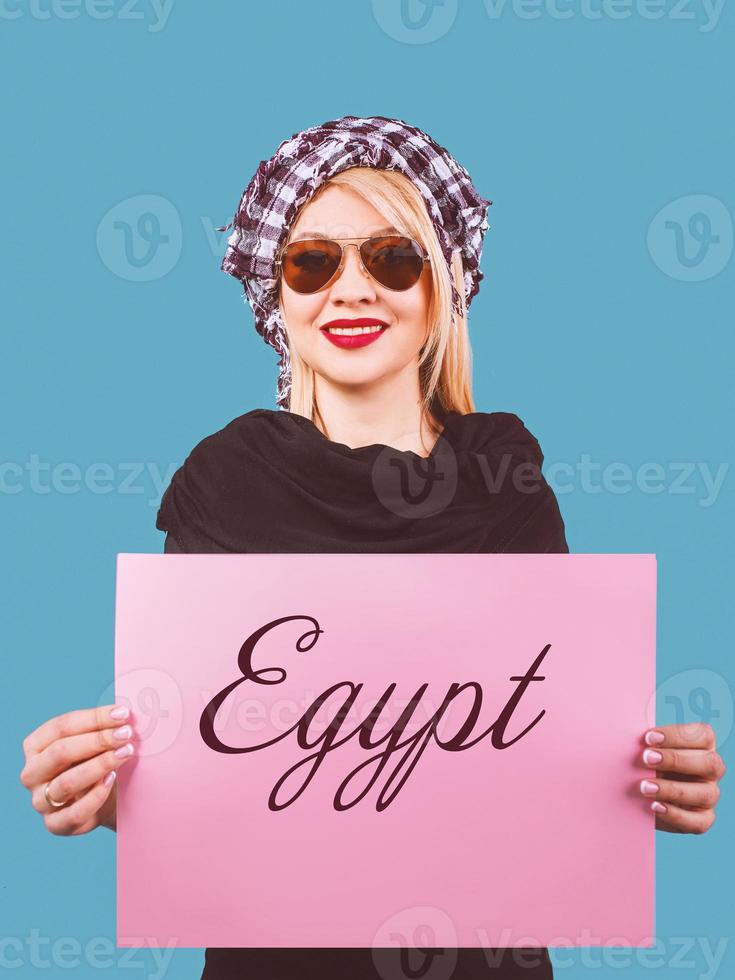 cute adorable cheerful smiling blonde woman in sunglasses and arafatka with pink empty table in her hands on blue background. Travel photo