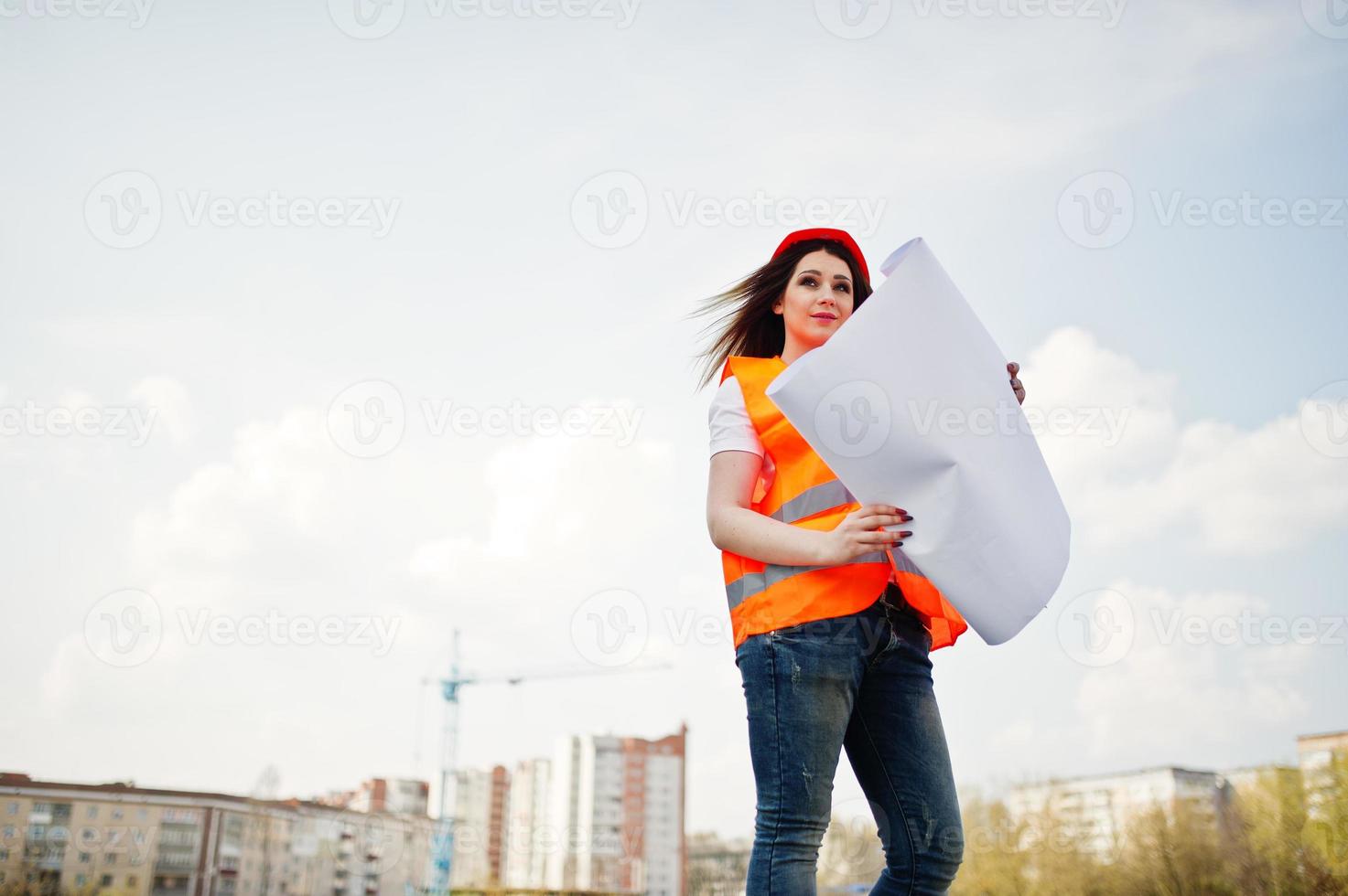 Engineer builder woman in uniform waistcoat and orange protective helmet hold business paper against new buildings with crane. photo